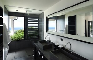 A modern bathroom with a large mirror, two sinks, and a view of greenery and the sea.