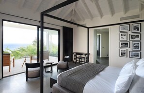 A modern bedroom with a large bed, a balcony with seating, and a view of the sea.