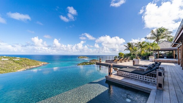 Une piscine à débordement luxueuse, avec vue sur une mer d’un bleu cristallin, agrémentée d’une terrasse en bois et de chaises longues.