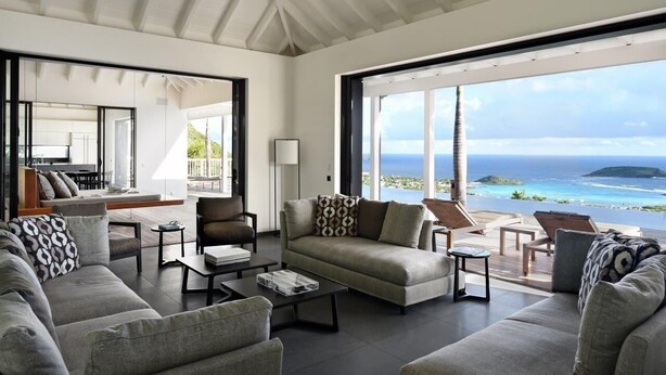 A spacious living room with a large sofa, armchairs, and a coffee table. There is a view of the ocean through large windows.