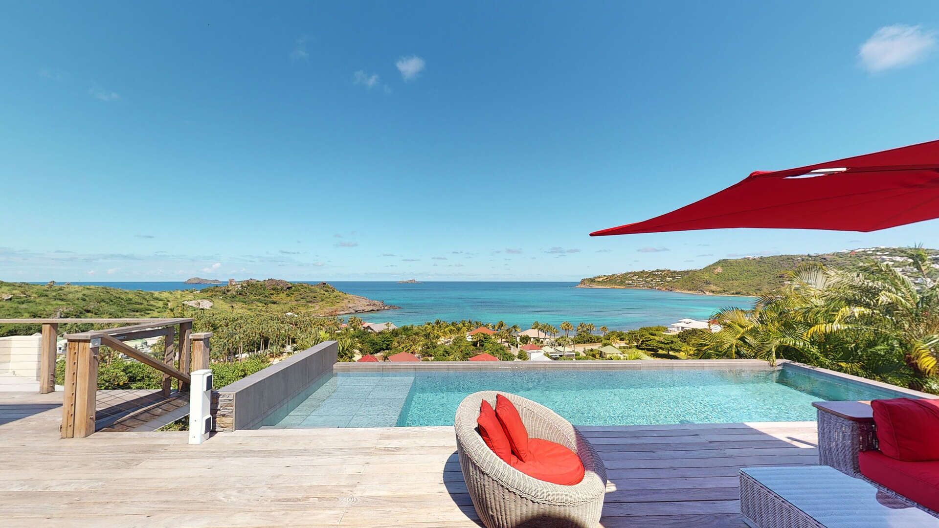 Zona de piscina con vista al océano, terraza de madera, sillas de mimbre y un paraguas rojo.