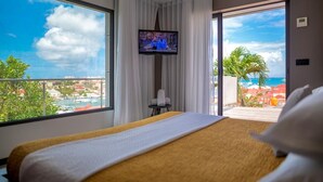 Ein Hotelzimmer mit einem großen Bett, einem Fernseher und einem Balkon mit Blick auf das Meer und Gebäude.