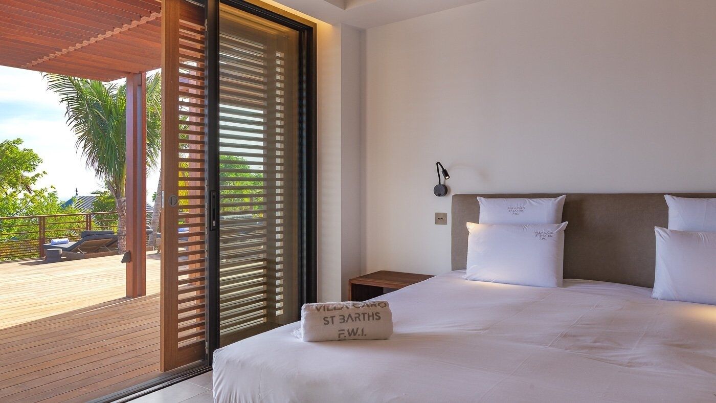A bedroom with a bed, pillows, a nightstand, and a view of a deck and palm trees.
