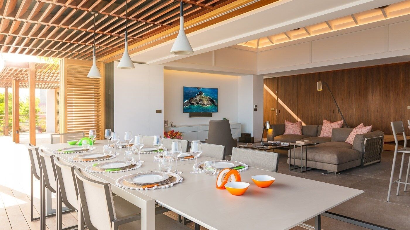 Un espace de repas moderne avec une longue table dressée pour six personnes, un canapé et un téléviseur en arrière-plan.