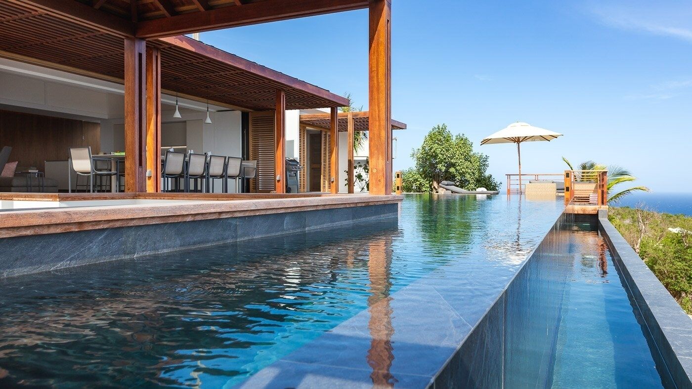 A modern outdoor pool area with a wooden pergola, seating, and a view of the ocean.