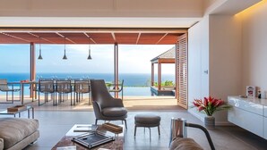 A modern living room with a view of the ocean, a dining area with a table and chairs, and a kitchen area with a white cabinet and a vase of flowers.