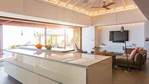 A modern kitchen with a large island, a dining area with a glass partition, and a living room with a sofa and TV.