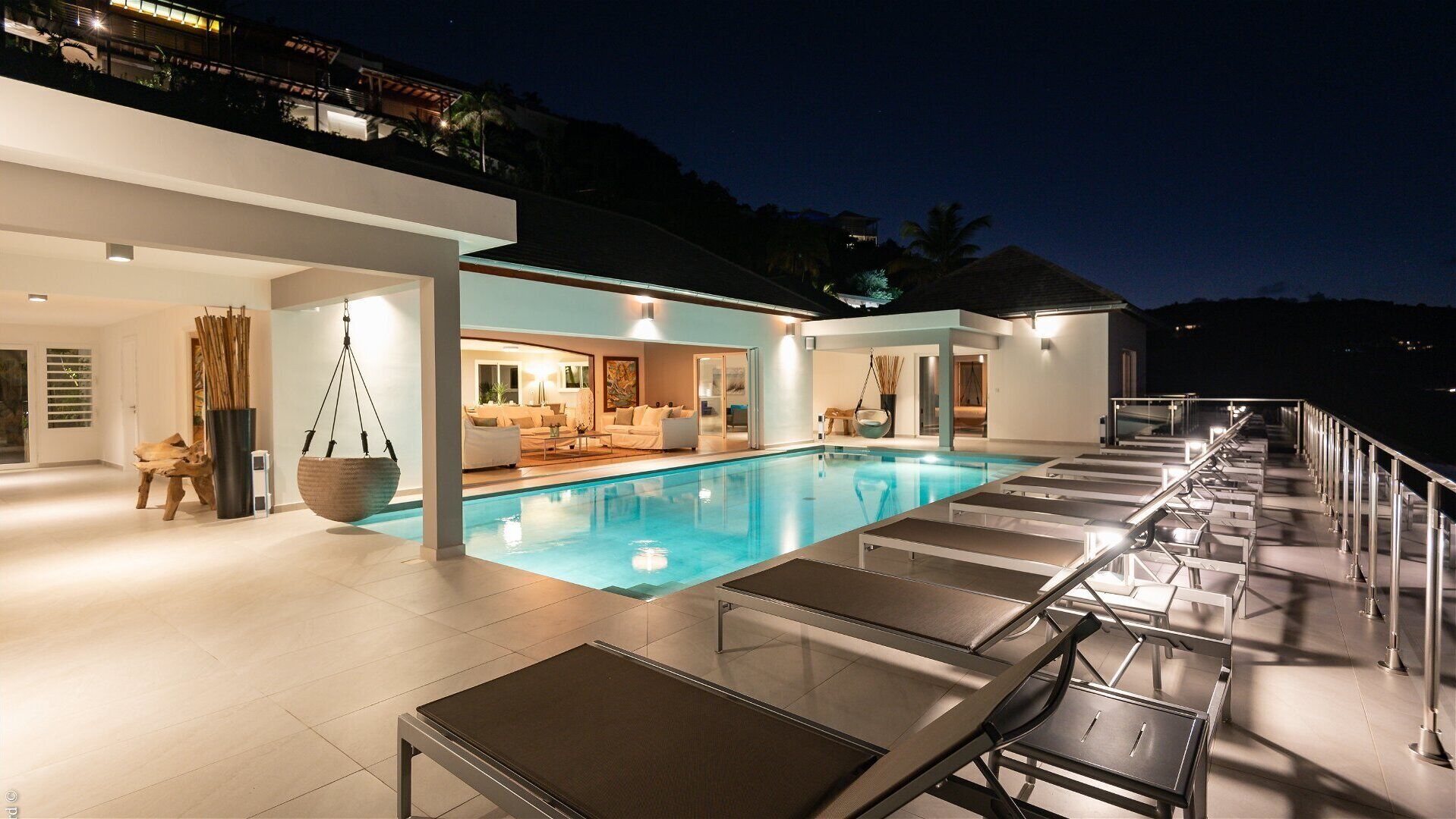 Un espace piscine extérieur moderne, doté de chaises longues, et un espace intérieur bien éclairé avec un canapé et un coin repas.