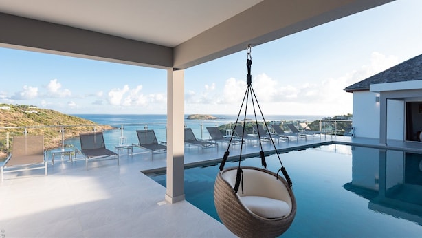 Ein moderner Infinity-Pool mit Hängesessel, Liegestühlen und Blick auf Meer und Hügel.