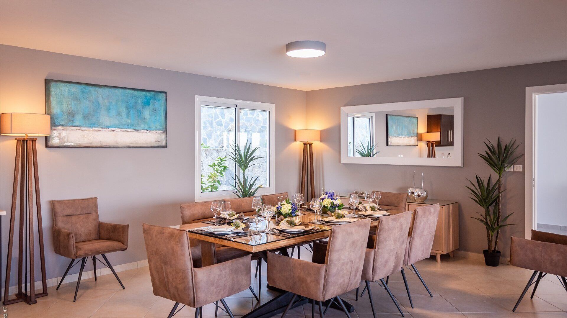 Uma sala de jantar com uma mesa retangular posta para seis pessoas, rodeada por seis cadeiras. Há duas luminárias de chão, uma janela grande com vista para o verde, e um espelho fixado na parede.
