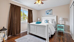 Une chambre avec un lit blanc, des rideaux marron, un ventilateur de plafond, une fenêtre avec vue, une table de chevet turquoise et une lampe blanche.