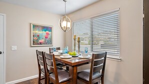Un coin repas avec une table en bois dressée pour quatre personnes, entourée de chaises. Un tableau orne le mur, un lustre est suspendu au-dessus de la table et une fenêtre est équipée de stores.
