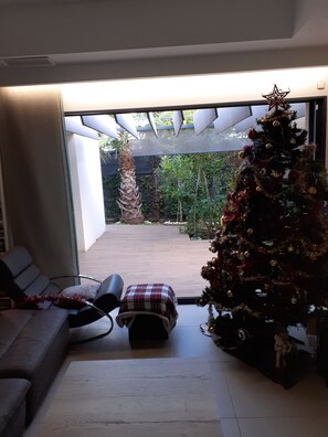 A modern living room with a large glass door leading to an outdoor patio. A Christmas tree is decorated with ornaments and placed near the door.
