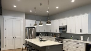 A modern kitchen with white cabinets, stainless steel appliances, a central island with stools, and pendant lighting. - Rare Find - Newer Construction close to town & beach! (Whitefish)
