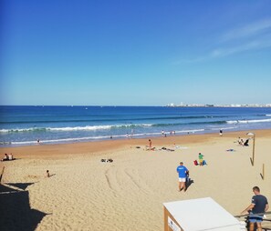 Una playa con gente tomando el sol y caminando por la orilla, cielo despejado y un mar tranquilo con barcos a lo lejos.