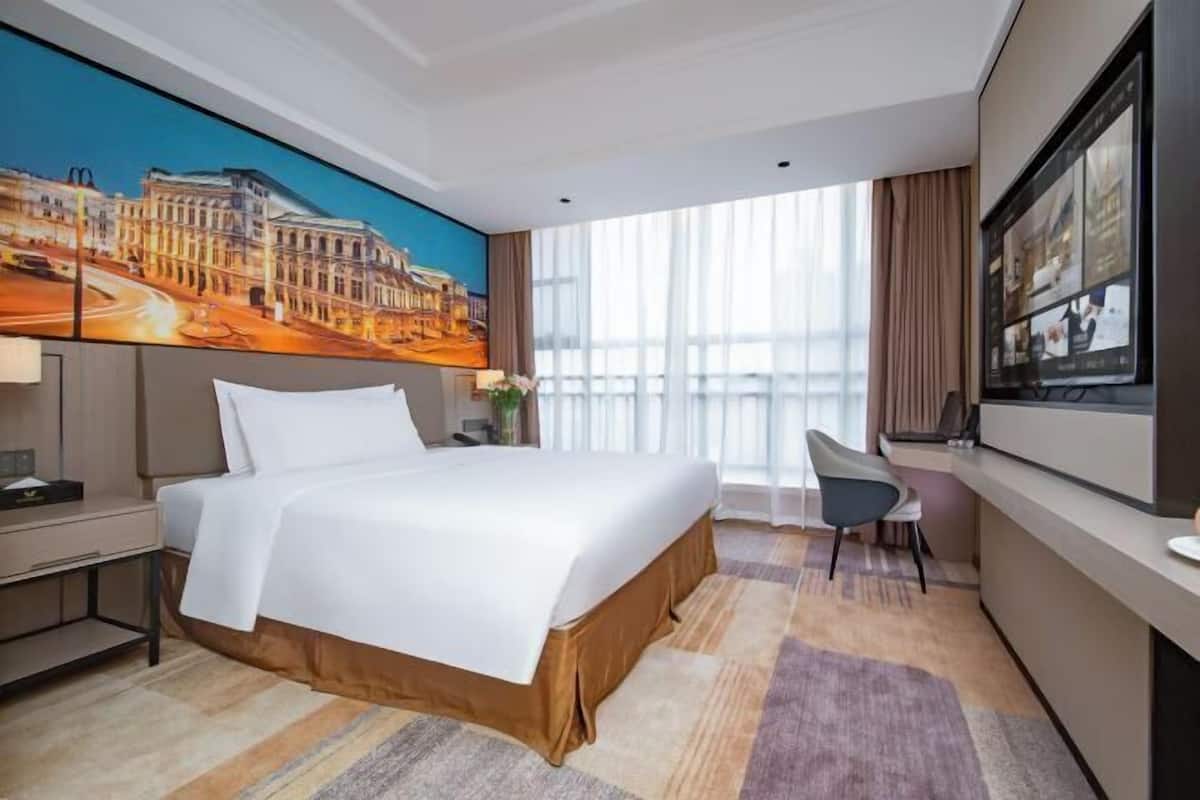 A hotel room with a large bed, a desk, a chair, a TV, and a large window with curtains.