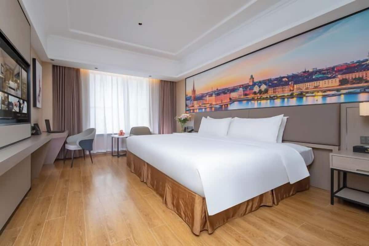 A hotel room with a large bed, a desk, a chair, a window with curtains, and a cityscape painting on the wall.