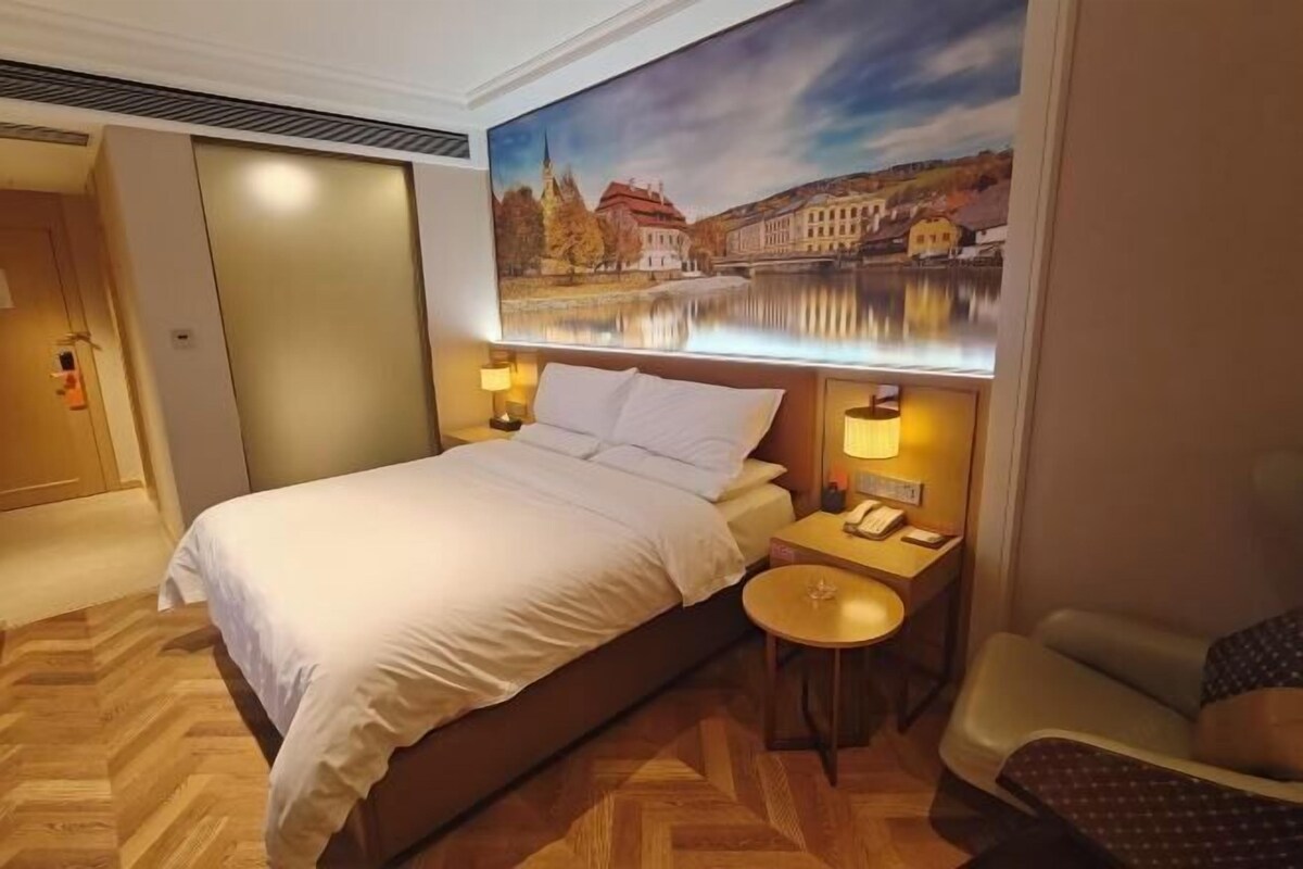 A hotel room with a bed, a nightstand with a lamp, a chair, and a painting of a scenic landscape.