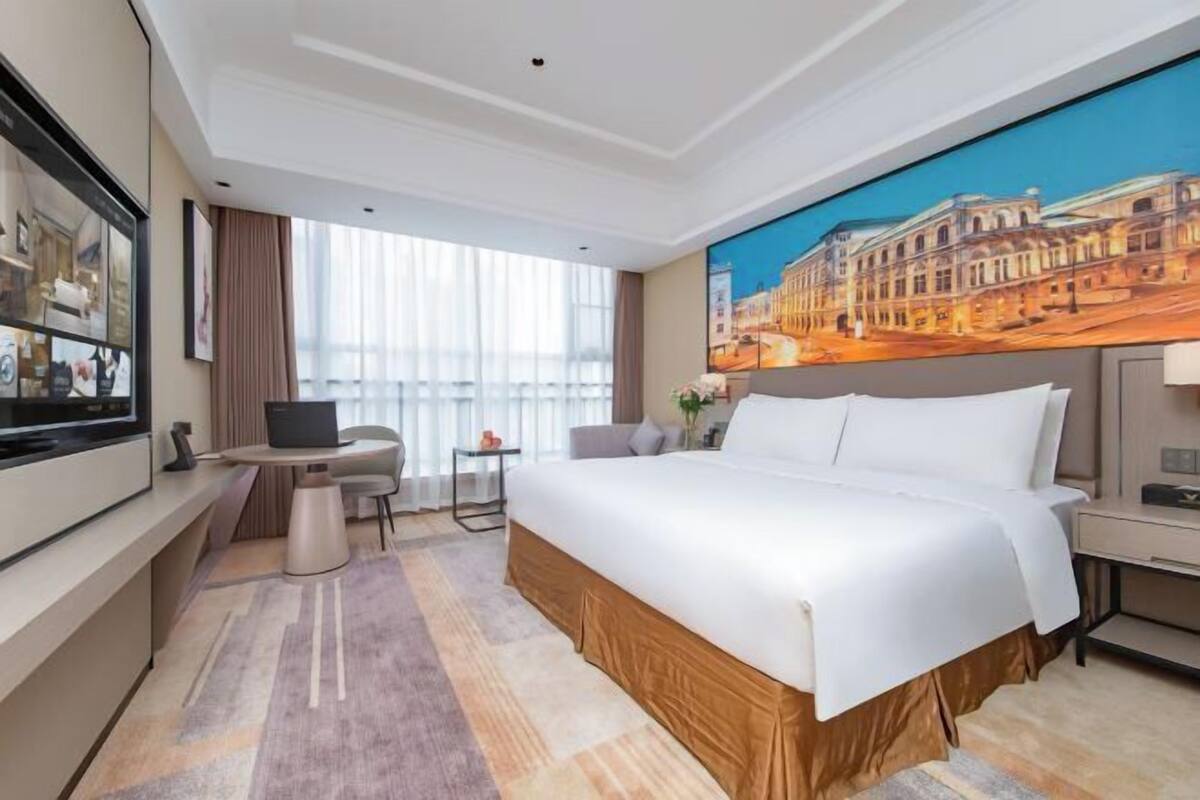 A hotel room with a large bed, a desk, a chair, a TV, and a painting of a cityscape.