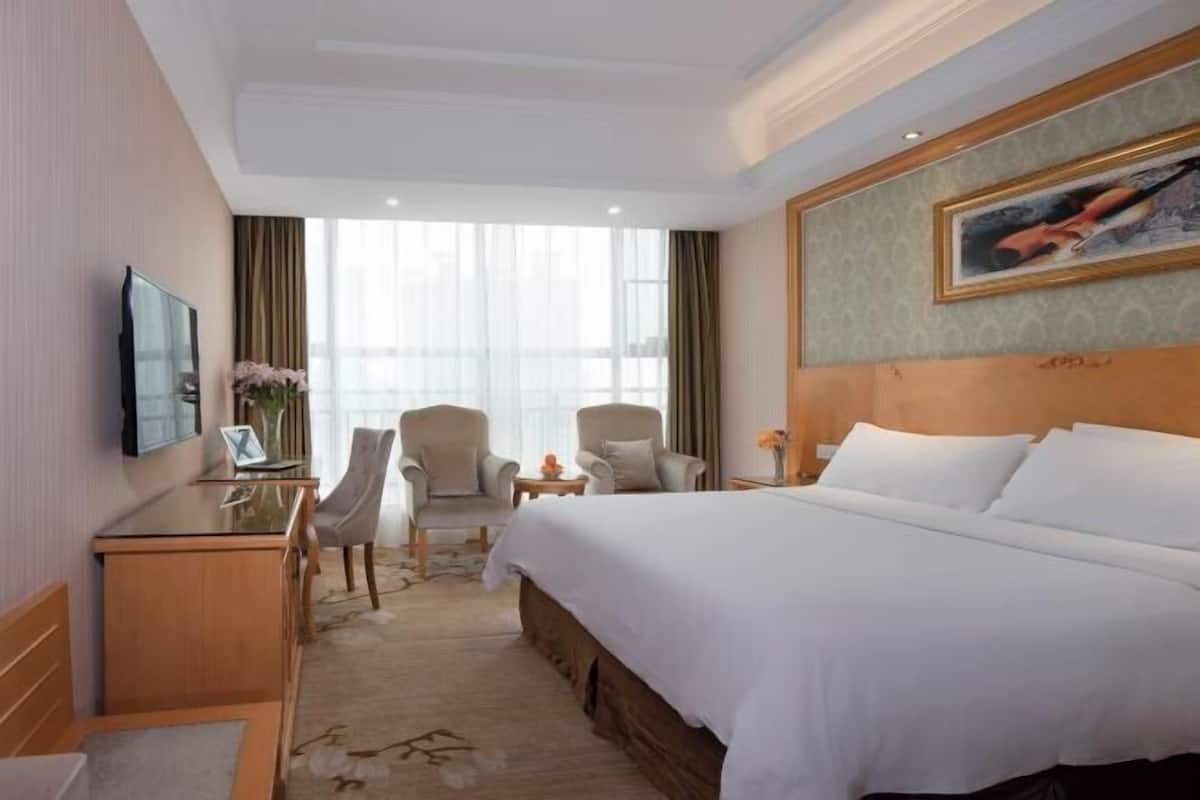 A hotel room with a large bed, a desk, two chairs, a TV, and a painting on the wall.