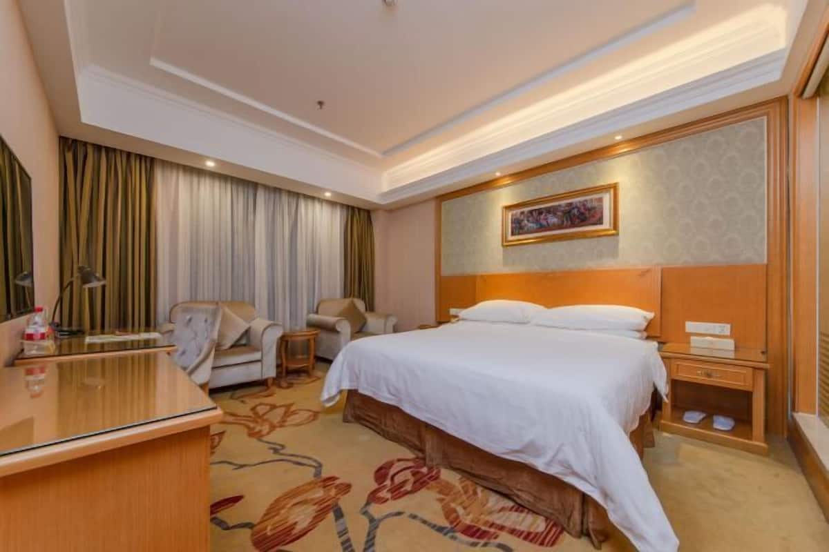 A hotel room with a bed, a desk, a chair, a nightstand, and a wall-mounted TV.