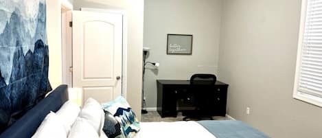 A bedroom with a bed, a desk, a chair, a door, a wall art, and a window.