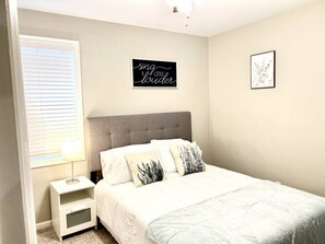 A bedroom with a bed, a nightstand, a wall art piece, and a window with blinds. - Vibrant 3BR Greensboro Home | Game Room & Yard (Greensboro)