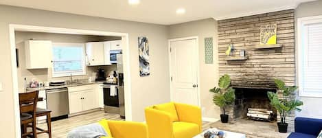 A living room with yellow armchairs, a white coffee table, a blue sofa, a fireplace, and a kitchen in the background.