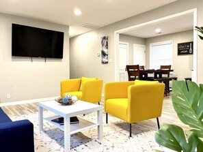 A modern living room with a flat-screen TV, yellow armchairs, a white coffee table, and a dining area with wooden furniture. - Vibrant 3BR Greensboro Home | Game Room & Yard (Greensboro)