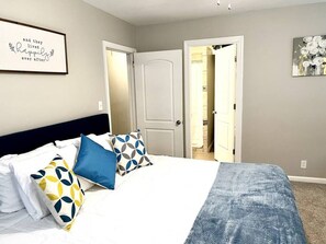 Un dormitorio con una cama, almohadas, una cita enmarcada en la pared y una puerta que da a otra habitación.