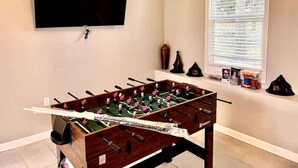 A room with a foosball table, a flat-screen TV mounted on the wall, and a window with blinds.