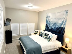 A bedroom with a bed, a TV mounted on the wall, a desk, and a large painting.