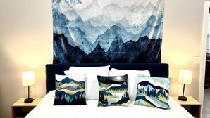 A bed with a large tapestry depicting mountains and a night sky, flanked by two bedside lamps and three decorative pillows with mountain and forest designs.