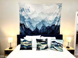 Un lit agrémenté d’une grande tapisserie représentant des montagnes et un ciel nocturne, flanqué de deux lampes de chevet et de trois coussins décoratifs aux motifs de montagnes et de forêts.