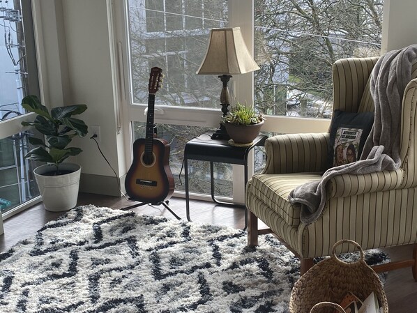 A cozy room with a large window, a guitar, a lamp, a potted plant, a striped armchair, and a woven basket.