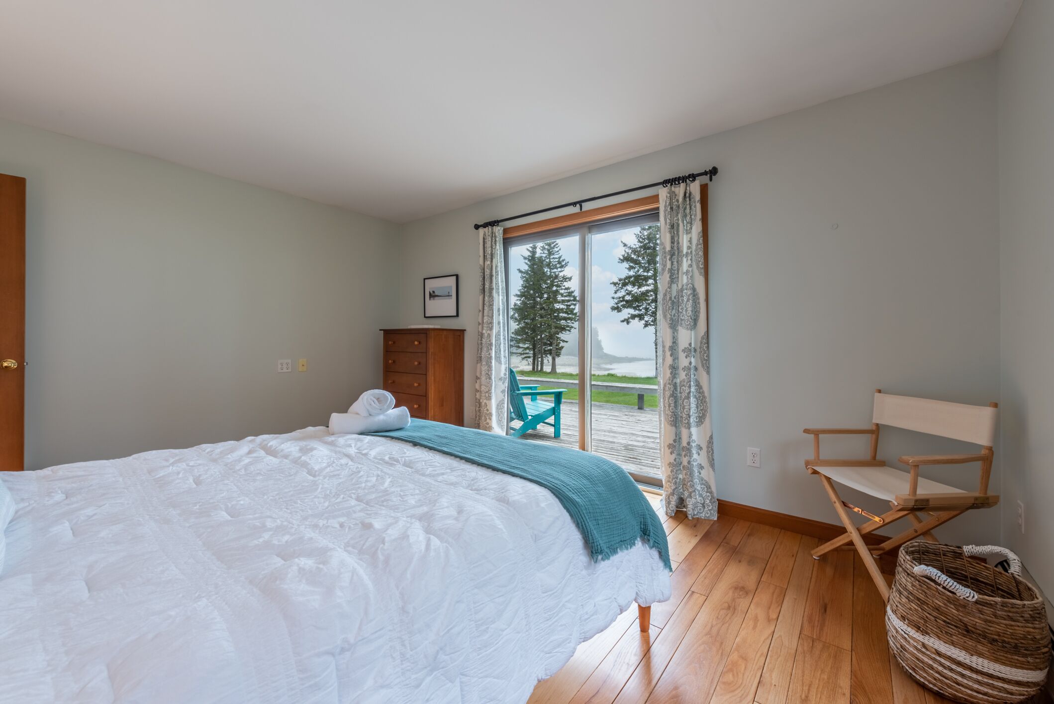 Une chambre avec un grand lit, une chaise en bois, un panier en osier et une vue sur une terrasse et des arbres, visible par une fenêtre.