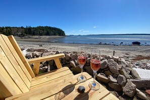 Ein Holzhocker mit zwei Gläsern Rotwein auf einem Tisch an einem felsigen Strand, mit freiem Blick auf den Ozean und die entfernten Hügel.