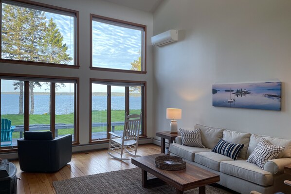 A spacious living room with large windows offering a view of a lake and trees, a comfortable sofa, a rocking chair, and a painting of a lake scene.