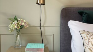 A wooden nightstand with a book and a vase of flowers, a wall-mounted lamp, and a bed with pillows. - Contemporary Urban Apartment (London)