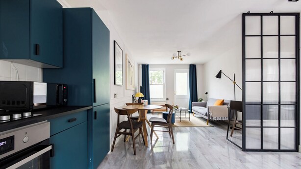A modern kitchen with teal cabinets, a dining area with wooden chairs, a sofa, and a large window with curtains. - Contemporary Urban Apartment (London)
