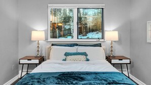Une chambre avec une grande fenêtre, deux lampes de chevet, un lit recouvert d’une couverture bleue et une vue sur un paysage enneigé.