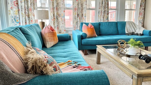 A living room with two blue sofas, patterned cushions, a glass coffee table, and floral curtains.