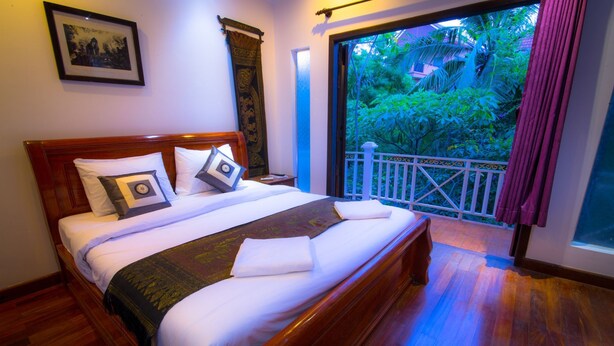 A neatly made bed with a wooden headboard, a balcony with a white railing, and a view of greenery outside. - Oasis Capsules - Hostel (Siem Reap)