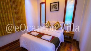 A neatly made bed with white linens and a decorative pillow, a wooden nightstand with a lamp, and a framed picture on the wall. - Oasis Capsules - Hostel (Siem Reap)