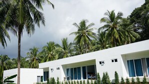 A modern white building with large windows and a balcony, surrounded by lush greenery and palm trees.