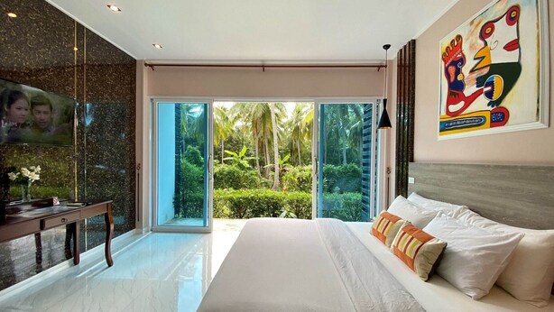 A modern hotel room with a large bed, a TV, a desk, and a sliding glass door leading to a tropical garden.