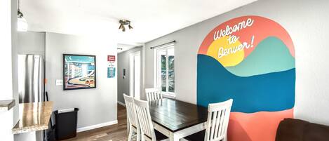A modern dining area with a wooden table and white chairs, a colorful wall mural, and a view of the outdoors.