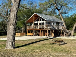 A two-story wooden cabin with a balcony, a covered porch, and a picnic table in front. - Treetop Oasis on the crystal clear Nueces River in the Texas Hill Country (UVALDE)