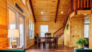 A spacious wooden interior with a high ceiling, a central dining area with chairs, and a staircase leading to an upper level.