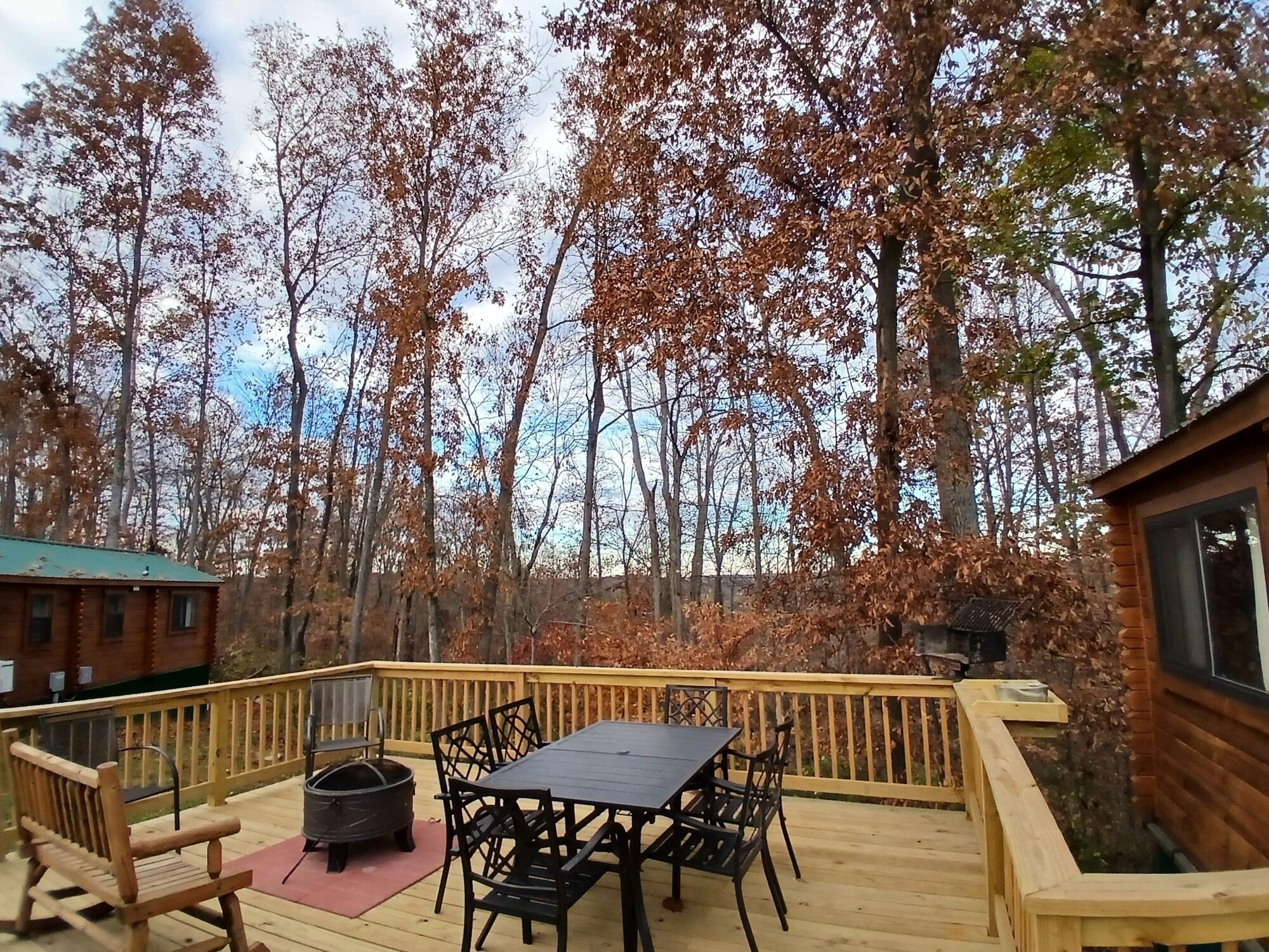A wooden deck with a table and chairs, a fire pit, and a view of a forested area.
